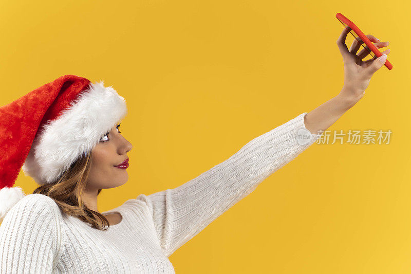 漂亮的年轻女子戴着圣诞老人帽，用手机/智能手机在黄色背景上视频通话/自拍